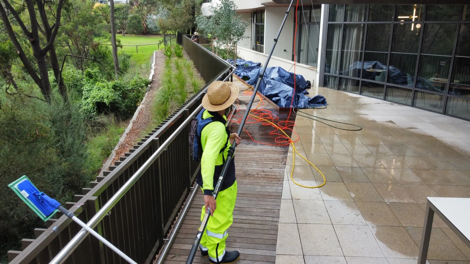 Technician in high-visibility gear using a water-fed pole to clean a modern commercial building exterior from a deck